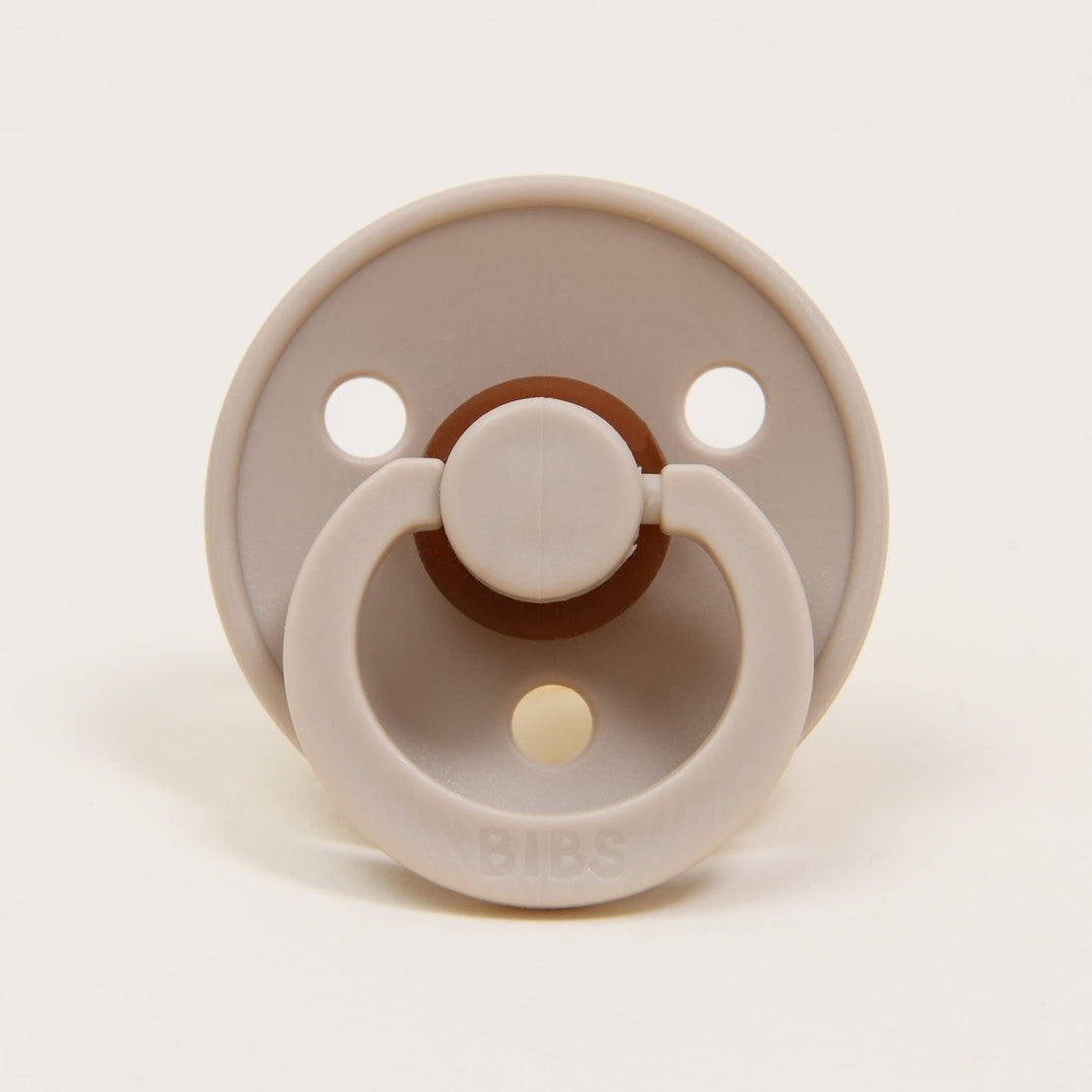 A Bibs Pacifier in "vanilla" color with three air holes, featuring a brown button handle and a ring, branded "bibs" on a natural background.