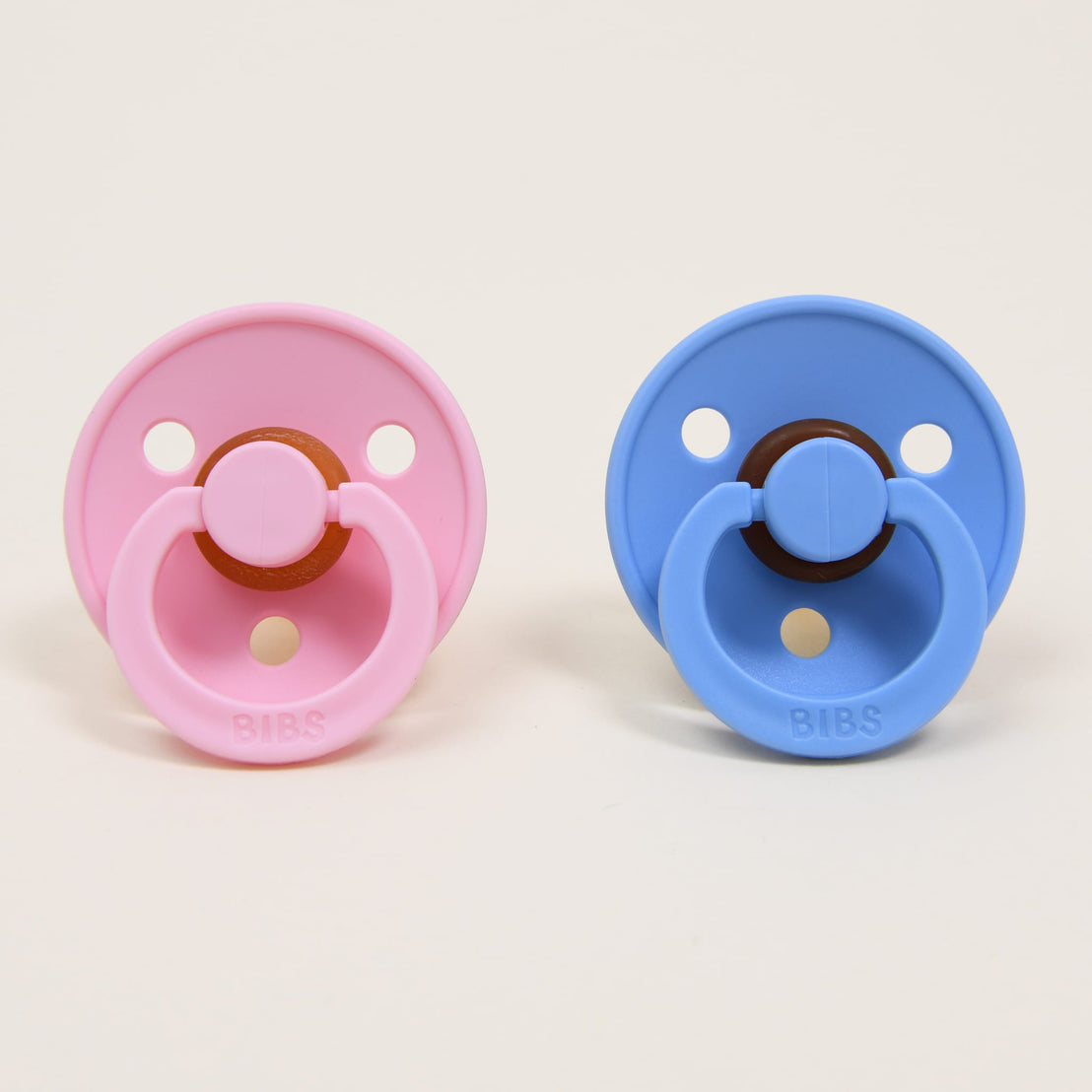 Two "Bibs" Pacifiers, one in "baby pink" and the other in "sky blue placed side by side against a beige background, each featuring a natural round button-style handle, perfect as an upscale baby gift.
