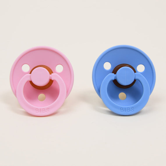 Two "Bibs" Pacifiers, one in "baby pink" and the other in "sky blue placed side by side against a beige background, each featuring a natural round button-style handle, perfect as an upscale baby gift.