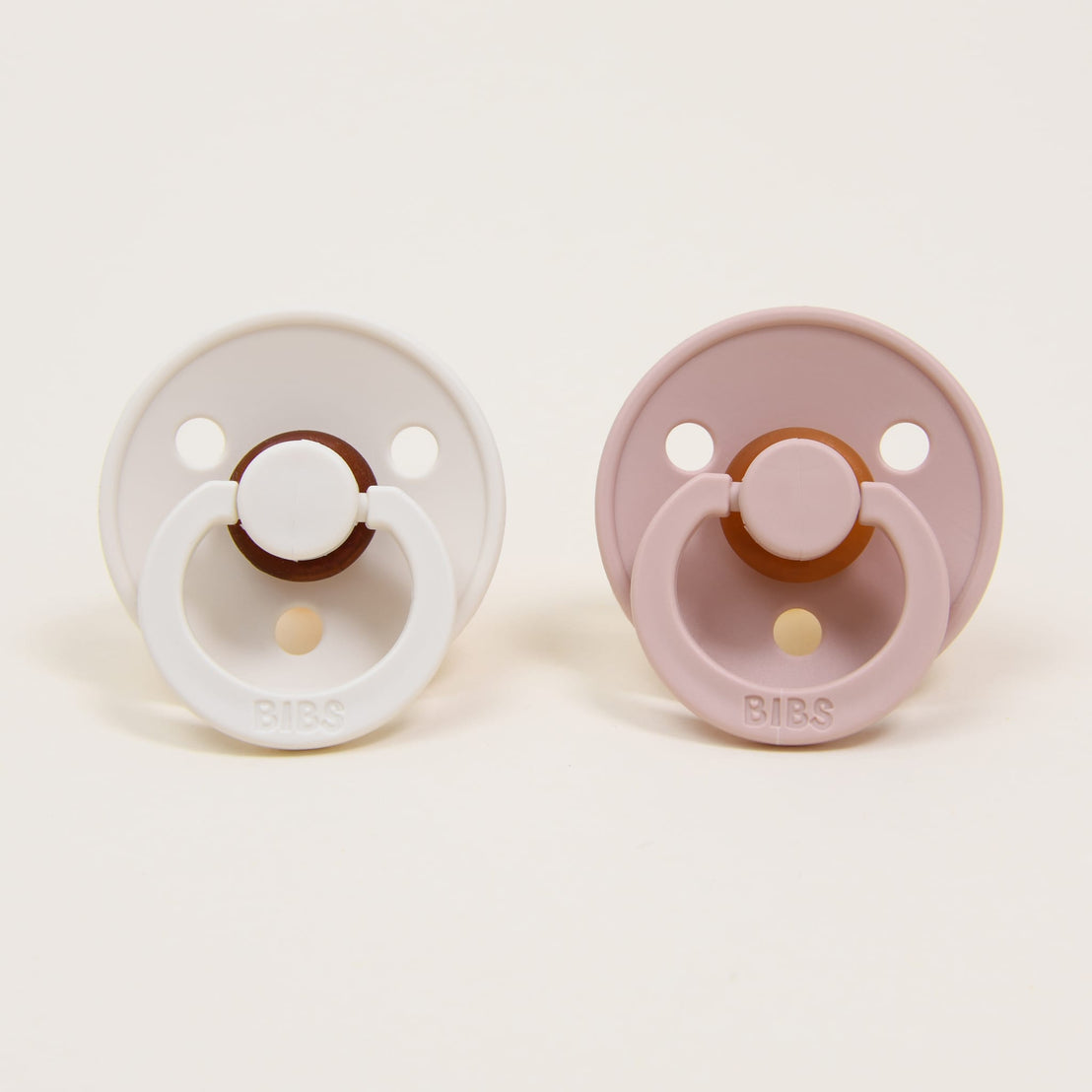 Two Bibs Pacifiers placed on a white background, one white and one mauve, both with round handles and three ventilation holes.