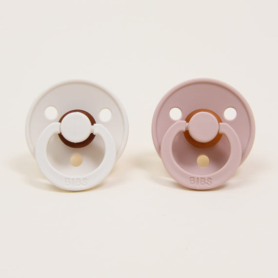 Two Bibs Pacifiers placed on a white background, one white and one mauve, both with round handles and three ventilation holes.
