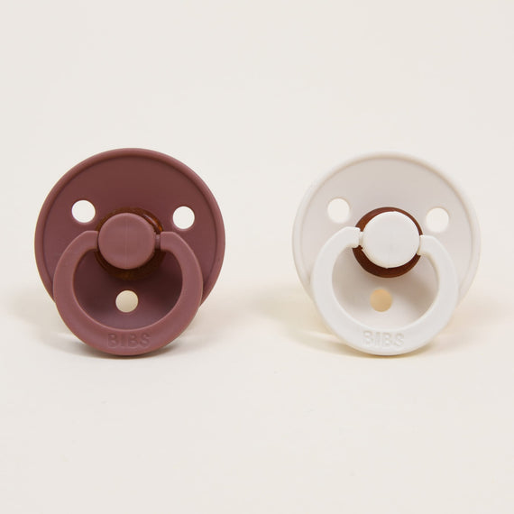 Two Bibs Pacifier Sets, one Woodchuck and one Ivory, displayed on a light background. Each upscale pacifier features a circular shape with three holes and a handle.