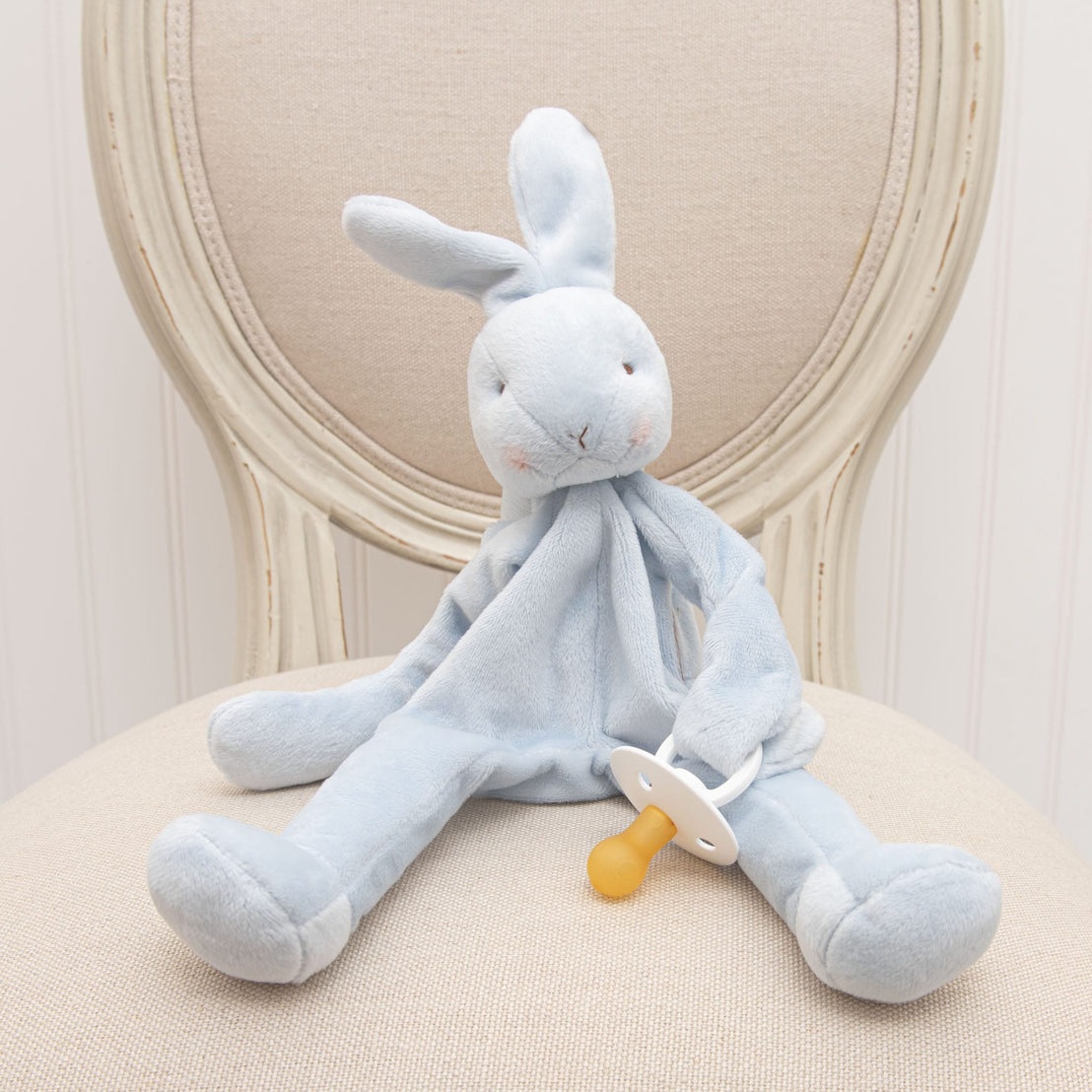 A Blue Silly Bunny Buddy | Pacifier Holder, an upscale baby gift, with a natural white nose sits on a cream upholstered chair. The bunny has long floppy ears and a serene expression.
