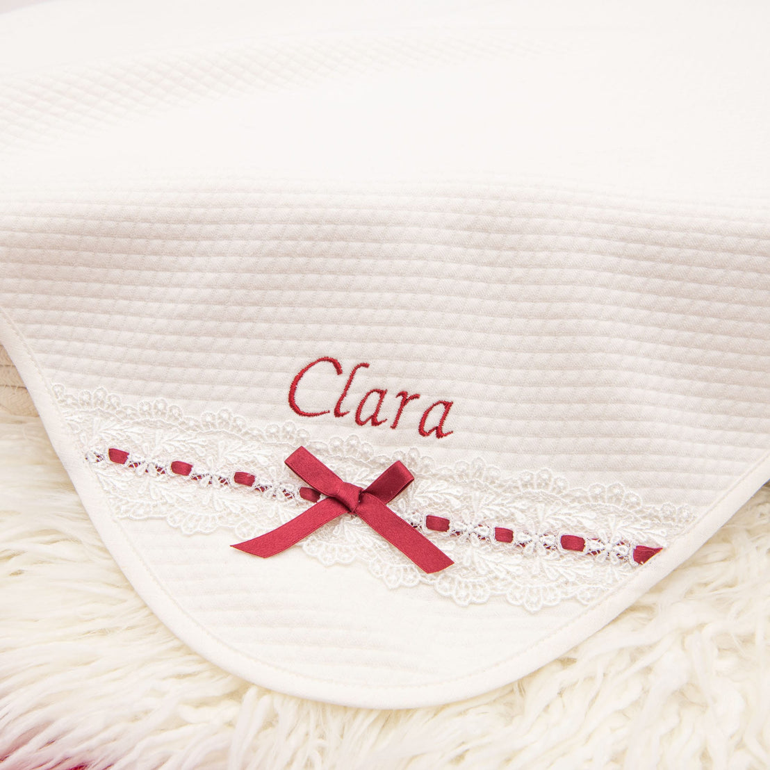 Introducing the Clara Gift Set - Save 10%: This luxurious, handmade-in-the-USA blanket features "Clara" embroidered in red on a soft white fabric. The blanket is enhanced with a beautiful lace trim adorned with a red ribbon and bow, all elegantly draped over plush white fur for an incredibly cozy and delicate look.