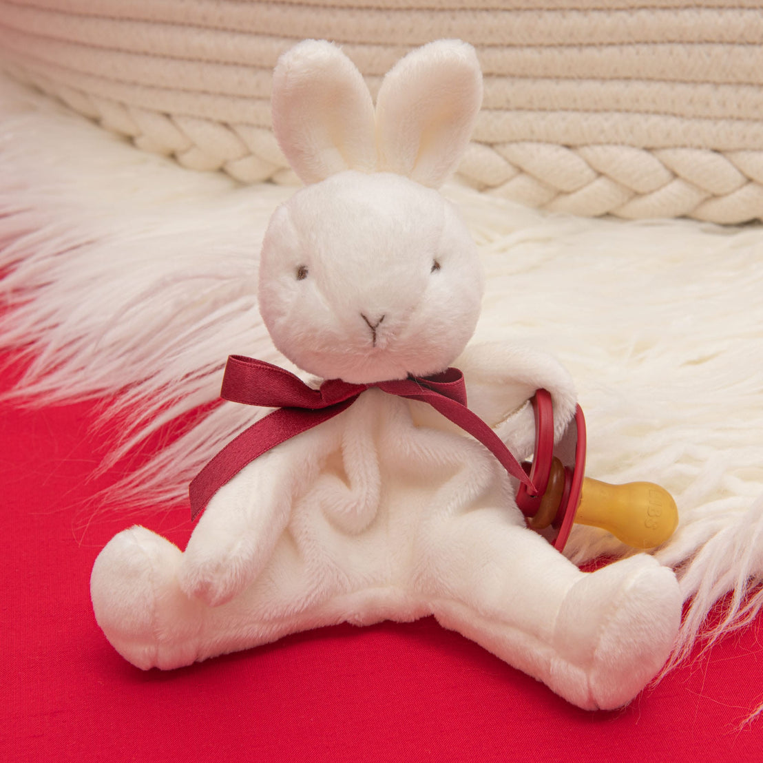 The Clara Gift Set - Save 10% features a cozy scene: a plush white bunny adorned with a red ribbon around its neck, sitting atop a quilted cotton surface. It clutches a pacifier while the backdrop showcases a braided basket and soft fur fabric, reminiscent of the comfort of a personalized blanket.