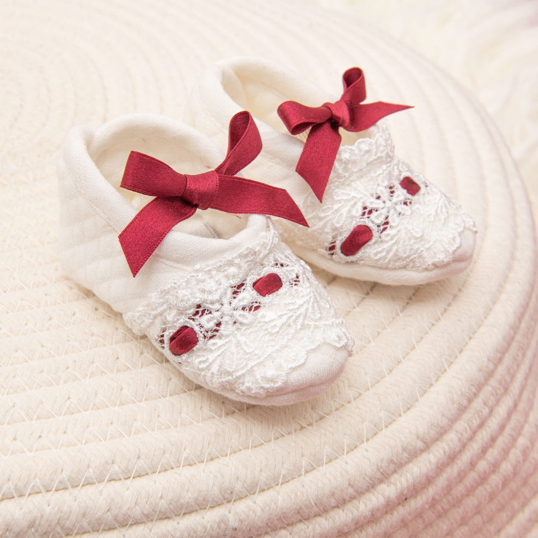 A pair of cute baby shoes from the Clara Gift Set - Save 10% rests on a textured surface, showcasing their white color with intricate lace details and red bows for a charming and delicate appearance reminiscent of handmade in USA craftsmanship.