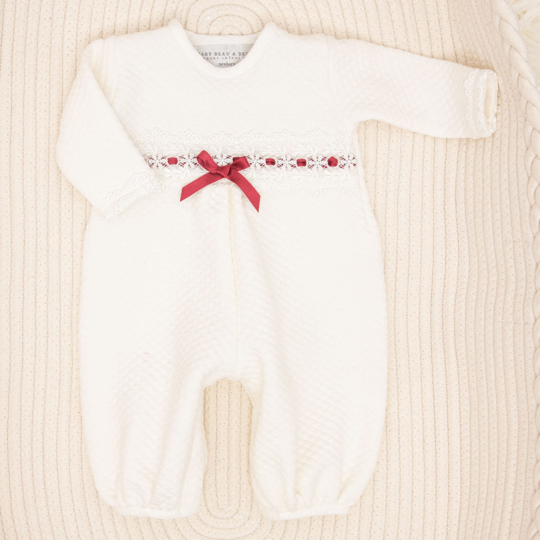 The Clara Gift Set, featuring a white baby romper adorned with lace details and a red ribbon bow across the chest, handcrafted from quilted cotton for maximum comfort, arranged on a textured beige surface, is currently available with a 10% discount.