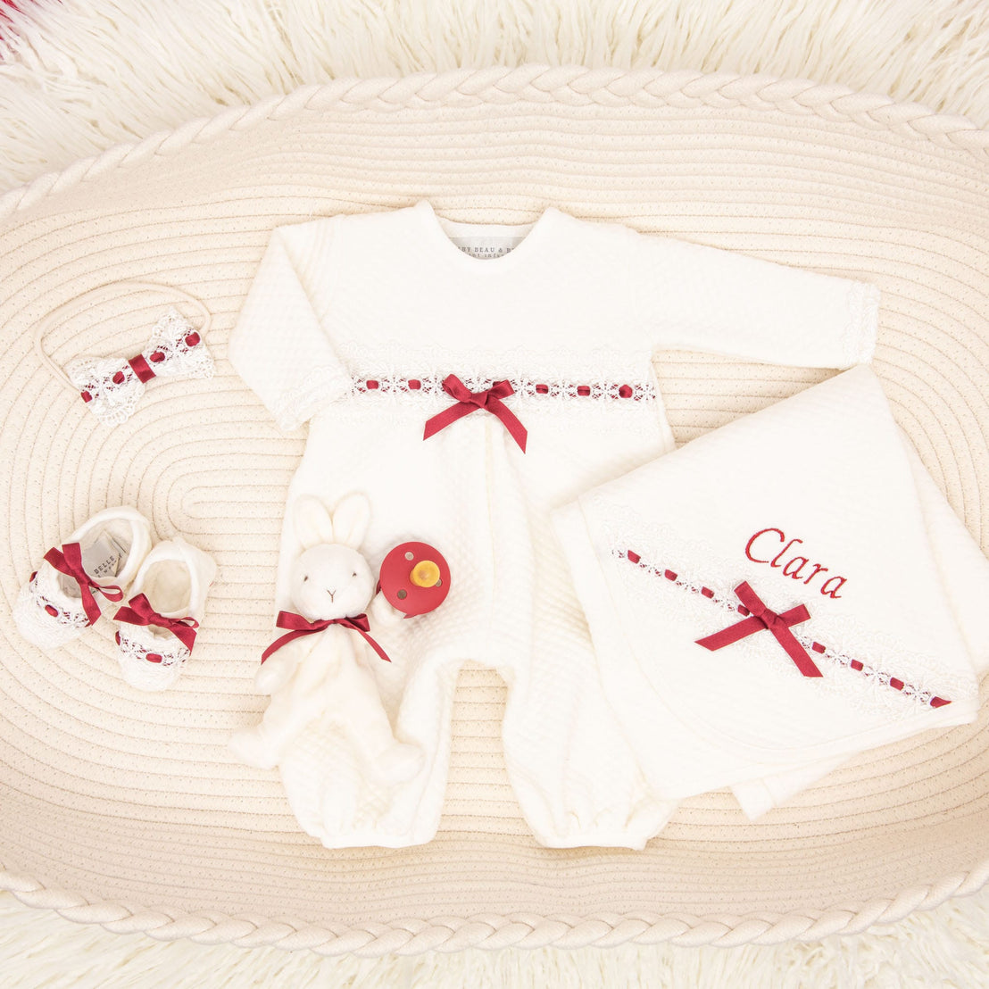 The Clara Gift Set - Save 10% features a delightful baby gift set that includes a white onesie adorned with red ribbons, coordinating shoes, a charming headband, a soft toy rabbit, and a red rattle. Crafted with care in the USA, this set is beautifully complemented by a personalized quilted cotton blanket with "Clara" embroidered in elegant red stitching. All items are artfully arranged on a textured cream rug for an exquisite presentation.