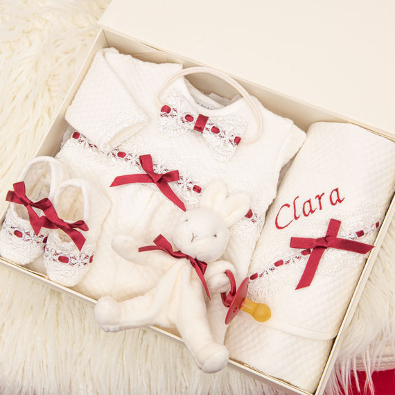 Introducing the Clara Gift Set - Save 10%: This charmingly arranged gift box includes a quilted cotton outfit embellished with lace and red bows, coordinating shoes, a cuddly white bunny toy crafted from handmade materials in the USA, and a personalized white blanket featuring "Clara" embroidered in red. All items are elegantly displayed on a fluffy white backdrop.