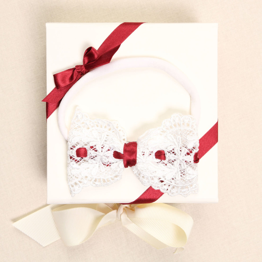A white gift box adorned with a red silk ribbon and bow, featuring an ivory lace and ribbon accessory topped with a red velvet bow, rests elegantly on a cream-colored surface, evoking the sophistication of the Clara Headband.