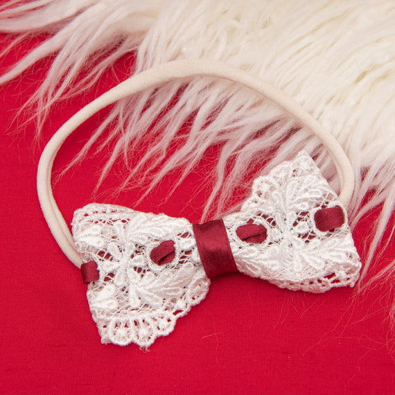 The Clara Headband is delicately crafted from ivory lace, adorned with maroon accents on a vibrant red silk ribbon, and elegantly displayed against a plush red fabric background with a white fur texture at the top.