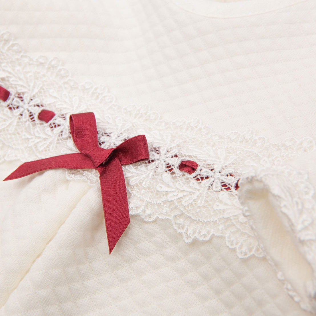 Close-up of the Clara Holiday Romper, a newborn jumpsuit made of quilted cotton, featuring detailed ivory lace trim and a small red bow. The lace showcases intricate patterns, while the bow adds a touch of contrasting color to the delicate design.