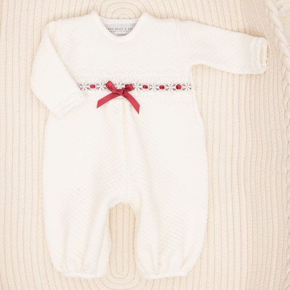 Introducing the Clara Holiday Romper: a cozy, white newborn jumpsuit featuring long sleeves and a textured pattern. It is beautifully embellished with delicate ivory lace detailing across the chest and adorned with a charming red ribbon bow. The romper is elegantly presented on a soft, cream-colored surface.