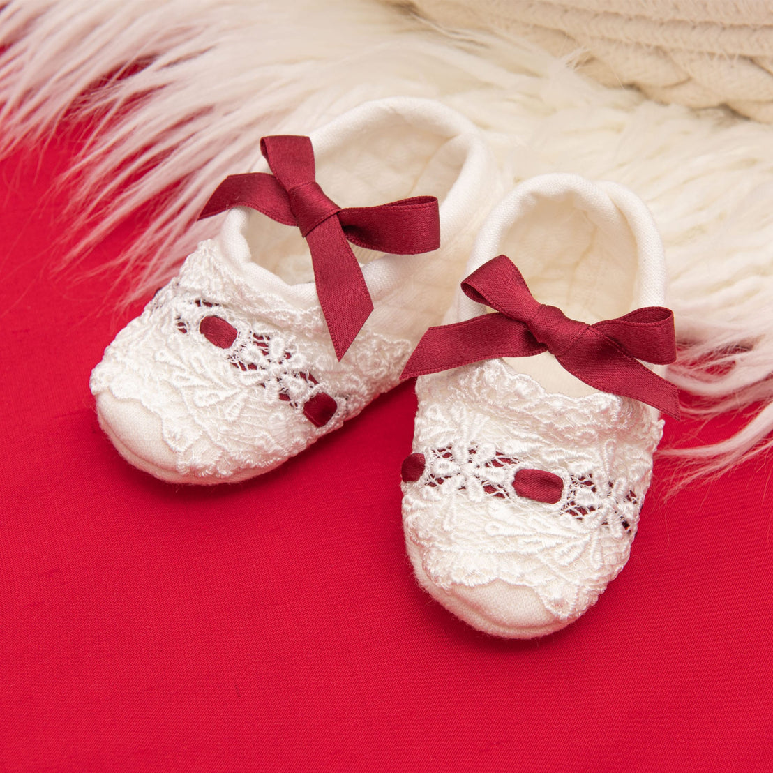 The Clara Quilted Booties, featuring lace detailing and maroon silk ribbon bows on top, rest on a plush white surface against a red fabric backdrop.