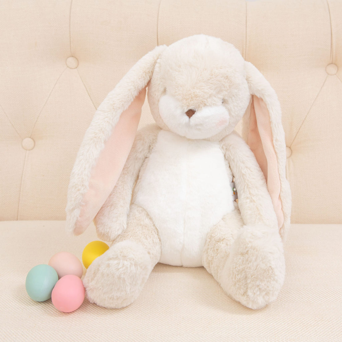 A Cream Nibble Bunny, specifically designed to be baby-safe and soft, sits on a beige tufted couch with its long ears. Next to it sit four pastel-colored eggs in pink, yellow, blue, and green, completing this adorable scene.