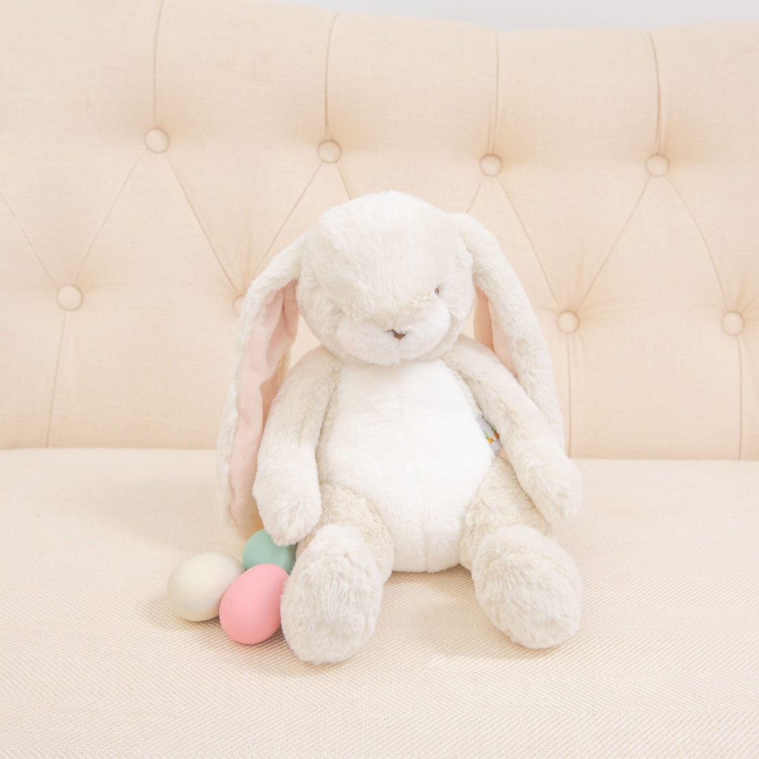 A Cream Nibble Bunny with long ears rests on a beige tufted couch, accompanied by three pastel-colored Easter eggs—pink, blue, and green. Baby safe and cozy, the setting is delightfully minimalistic.