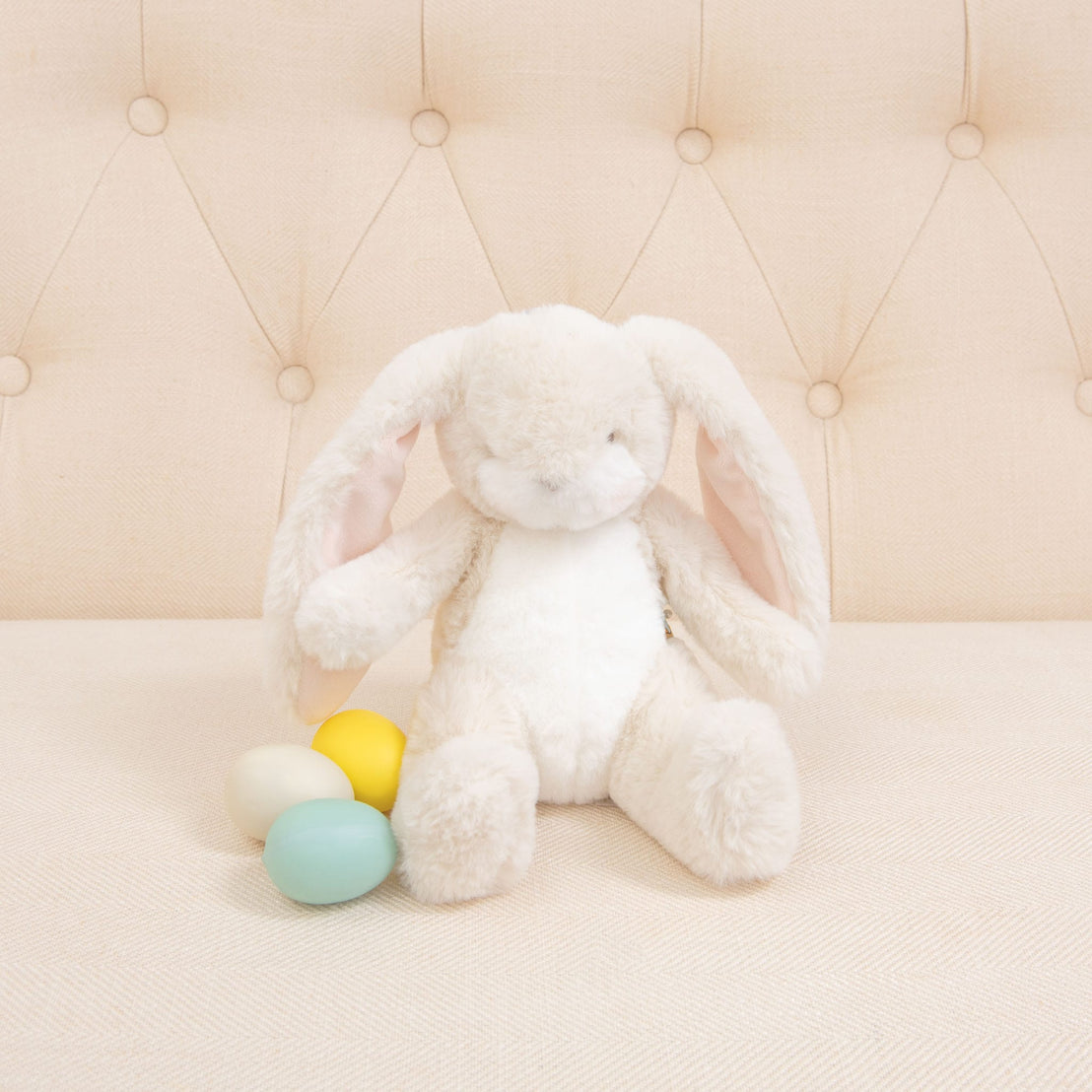 A Cream Nibble Bunny with long ears sits on a beige, tufted couch. Three pastel-colored eggs—yellow, blue, and cream—are nestled beside the baby-safe bunny.
