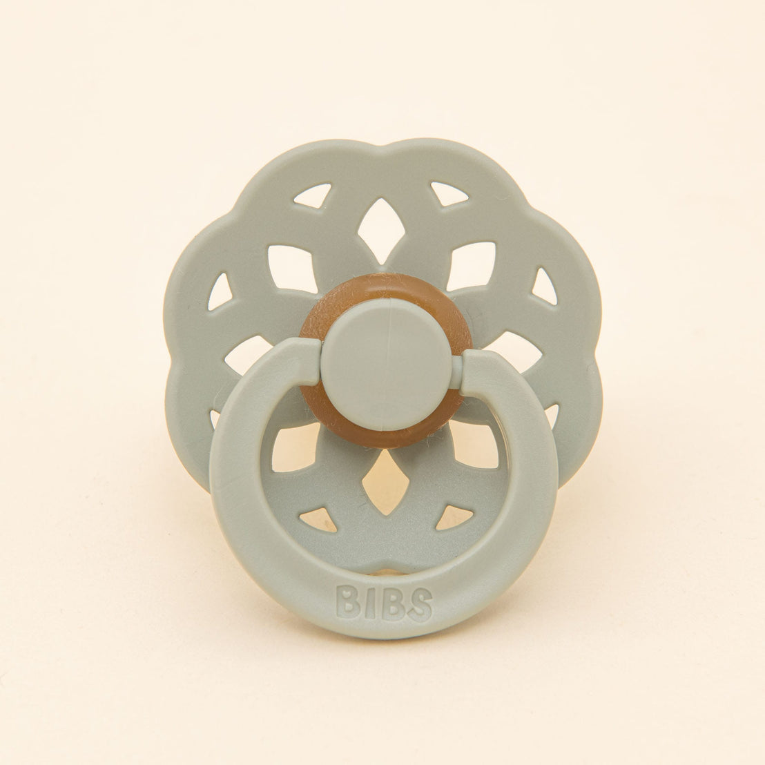 A Eliza Pacifier Set - baby pacifier with a round nipple and decorative, airy shield, styled as a flower for christening, on a light beige background.