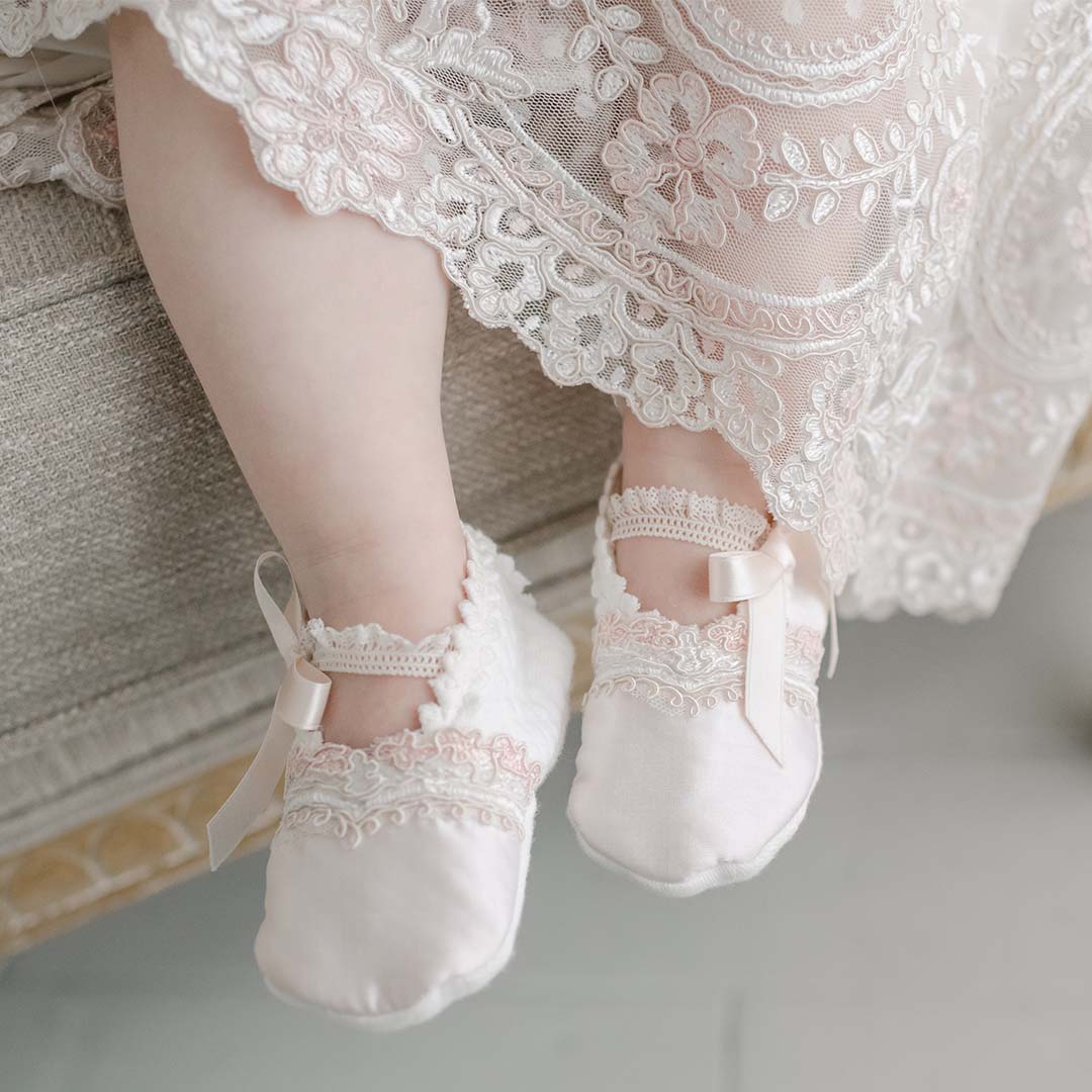 A close-up of a baby's feet wearing delicate Elizabeth Booties, possibly an heirloom, with satin ribbons, dangling in the air against a soft, pale background.