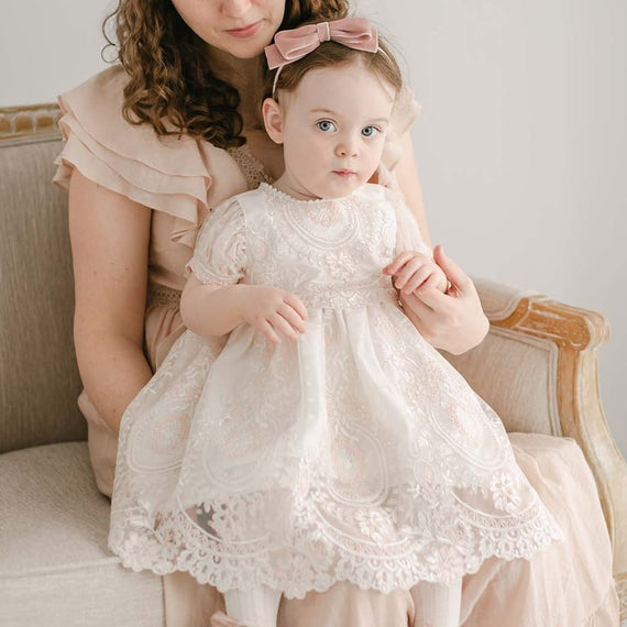 A woman with curly hair sits on a beige chair, holding a toddler in the Elizabeth Dress, which features embroidered lace. The toddler wears pink shoes and a pink bow headband. The woman gazes down at the child with a gentle expression.