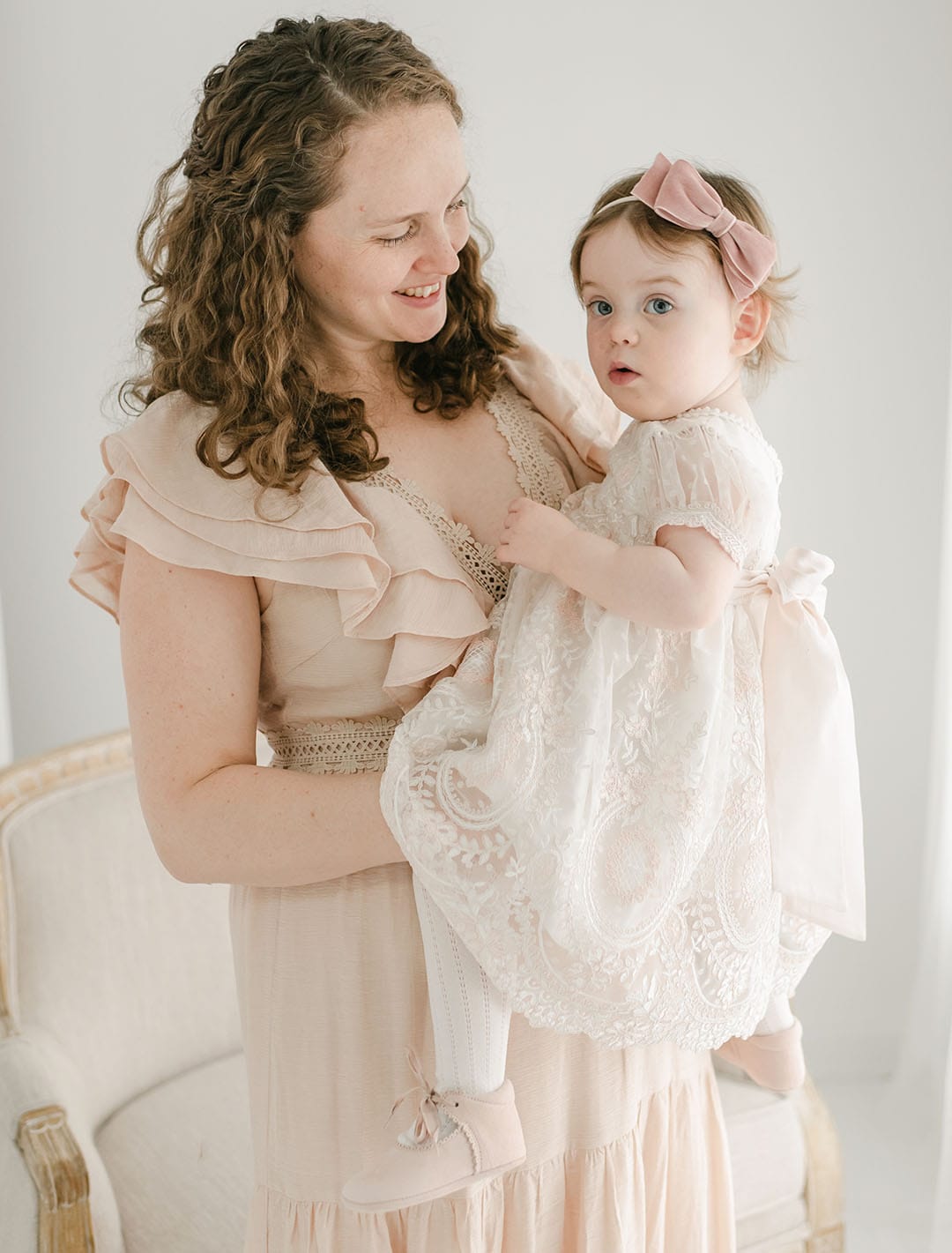 A woman with curly hair in a ruffled dress cradles a young child wearing the Elizabeth Dress, featuring embroidered lace and a delicate bow headband. They are in a softly lit room with light-colored furniture, both looking content.