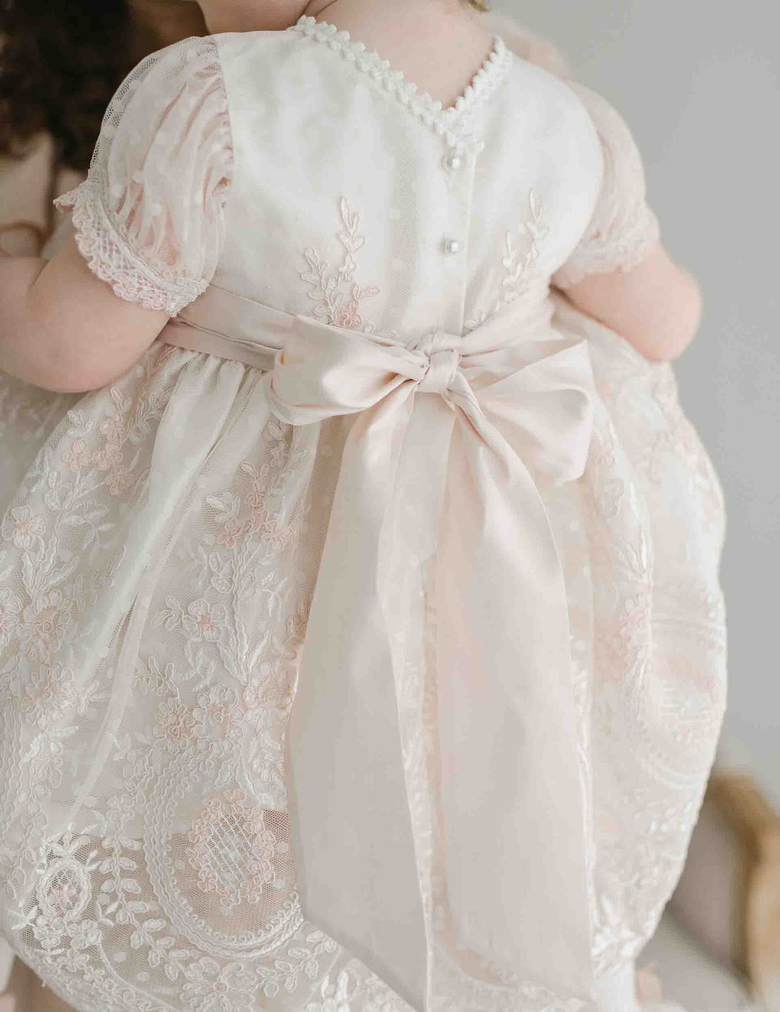 A baby wearing the elegant Elizabeth Dress showcases its intricate embroidered lace patterns and a large satin bow at the back. This cream-colored dress features delicate puffed sleeves and decorative buttons. Viewed from behind, it highlights a detailed design perfect for a baptism ceremony.