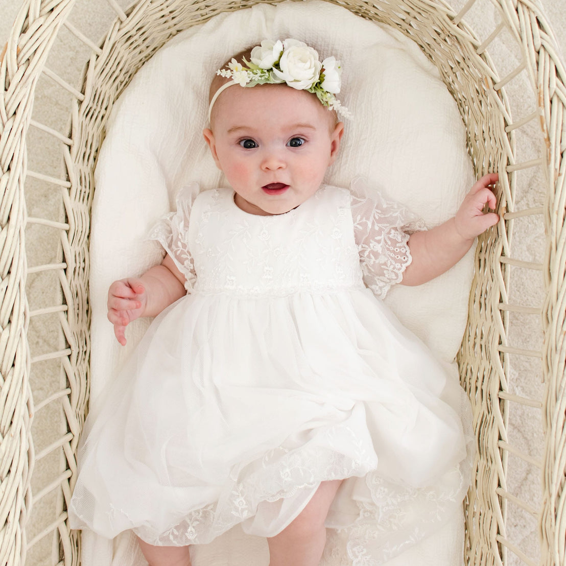 A baby wearing a vintage-inspired Ella baptism dress and flower headband lies in a wicker bassinet.