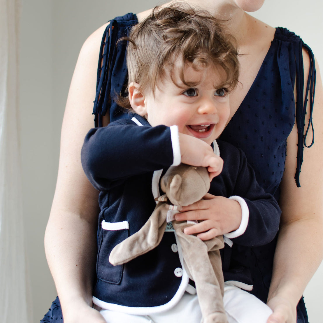 A smiling toddler with curly hair, wearing the handmade Elliott Jacket featuring a dark design with white trim, holds a stuffed bunny while sitting on an adult's lap. The adult wears a sleeveless navy French terry cotton dress, their heads not fully visible.