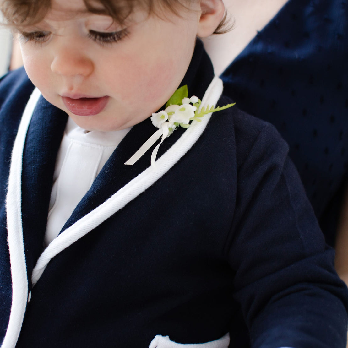 A toddler wearing the Elliott Jacket, a navy French terry cotton blazer with white trim, over a white shirt. The handmade jacket is adorned with a small boutonniere featuring white flowers and green leaves. The child is looking down, and part of an adult is visible in the background.