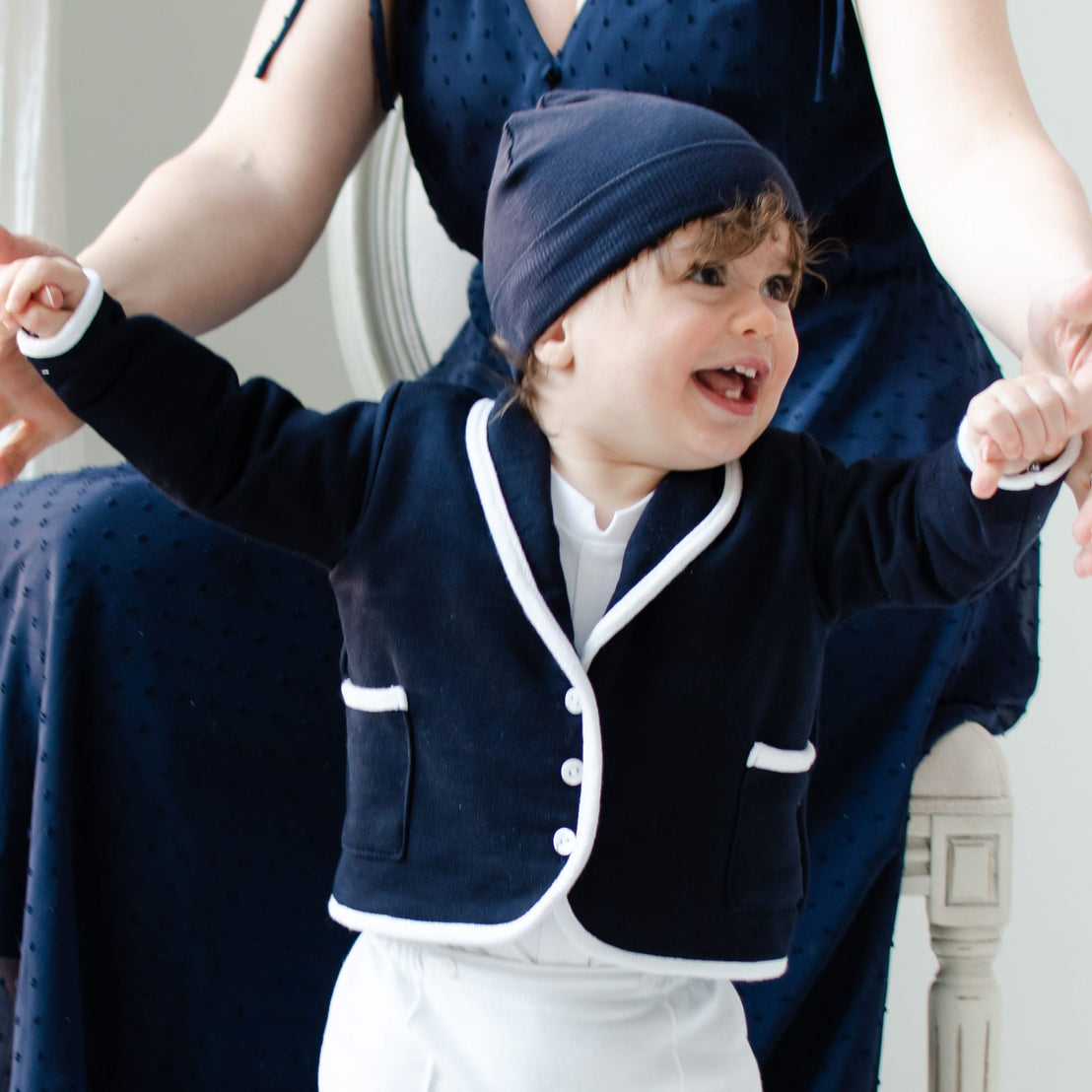 A joyful toddler wearing a navy Elliott Jacket with white trim holds onto someone's hands. The child smiles broadly while a person in a navy dress sits behind them, partially visible.