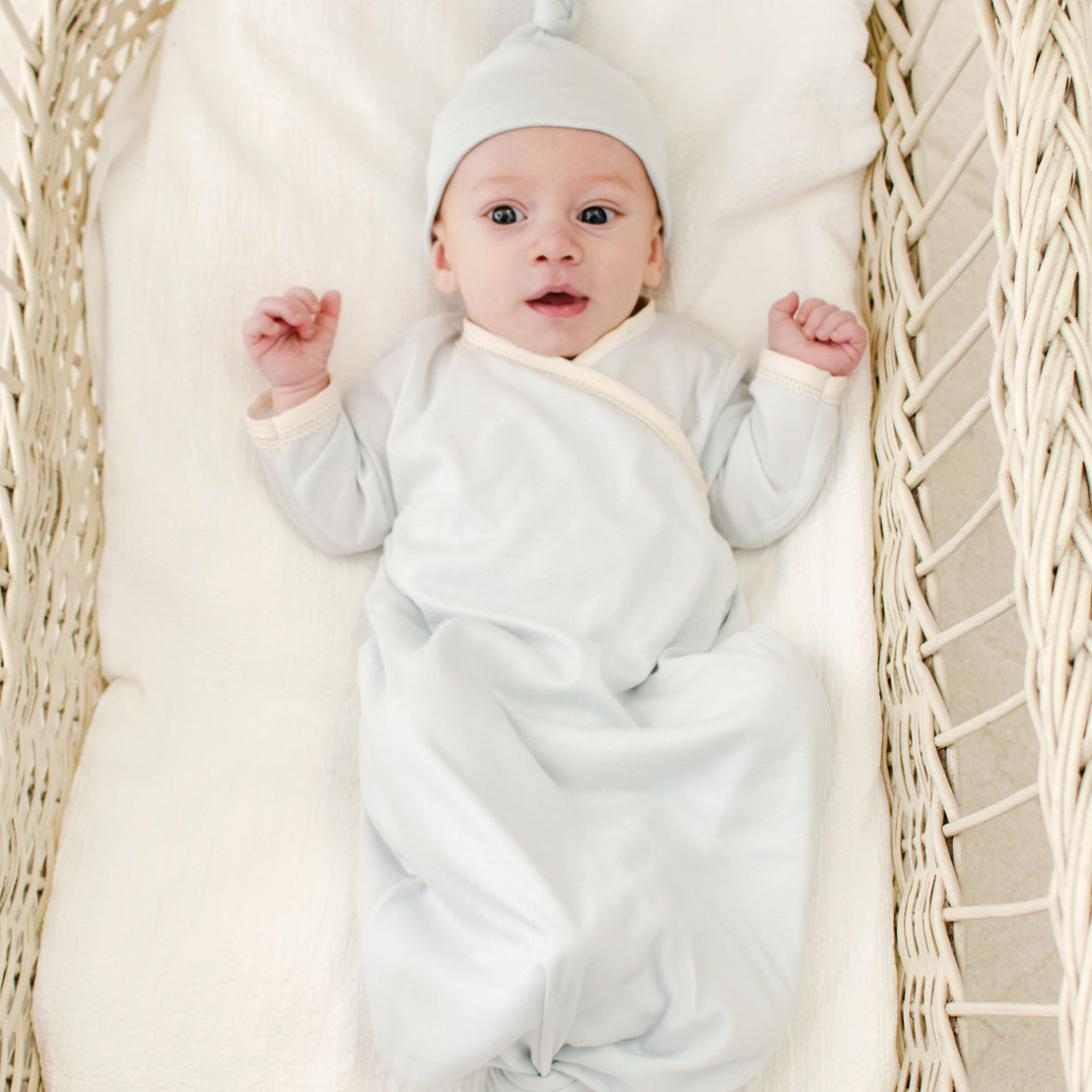 A baby in the Evan Knot Gown, made of 100% pima cotton, lies in a wicker bassinet on a soft white blanket, looking up with arms raised.