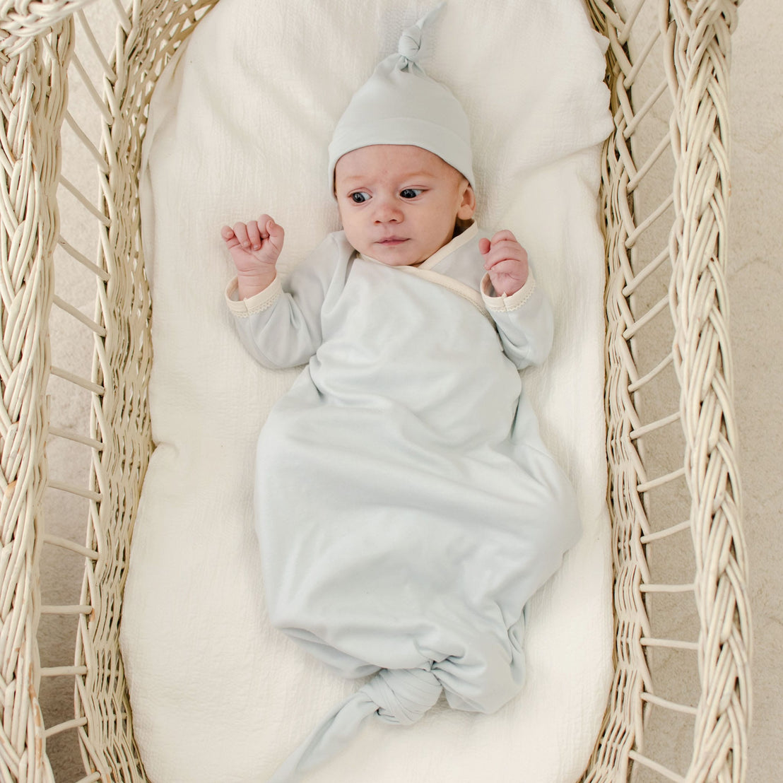 A newborn boy wearing an Evan Knot Gown, crafted from 100% pima cotton and handmade in the USA, rests quietly in a woven bassinet. The gown's soft, light hues complement the intricate patterns around him, creating a calming and gentle atmosphere.
