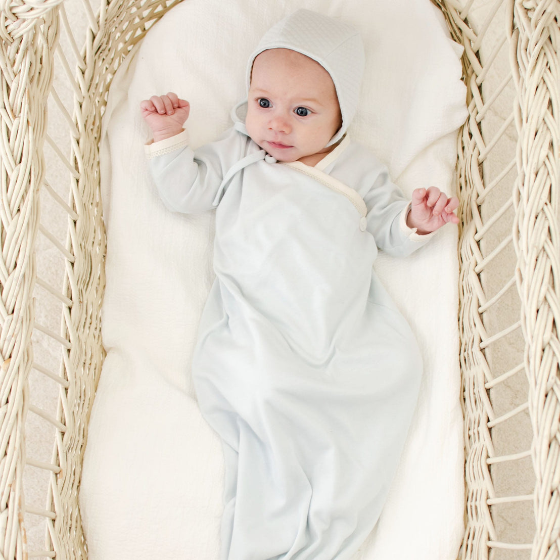An infant dressed in an Evan Knot Gown made from light gray 100% pima cotton, complete with a matching hat, is nestled comfortably in a woven bassinet. The baby gazes upward with gently raised arms, while the bassinet, equipped with a plush white cushion, exemplifies quality handmade craftsmanship from the USA.