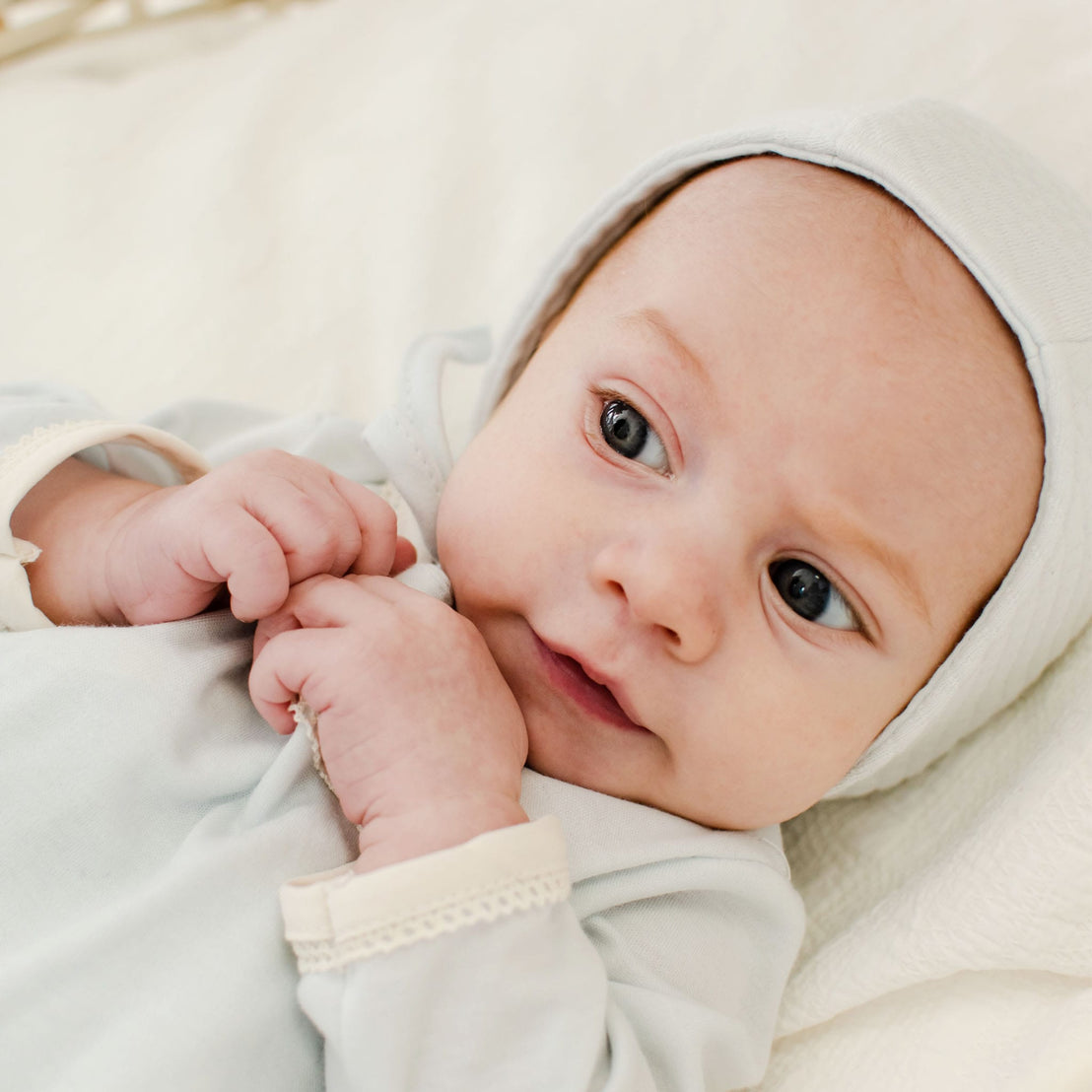 A baby wearing the Evan Knot Gown, crafted from 100% pima cotton in a soft white shade, is lying down and gazing upwards with wide eyes. The infant gently clutches the fabric and appears both relaxed and curious. The background showcases matching soft white fabric, all meticulously handmade in the USA.