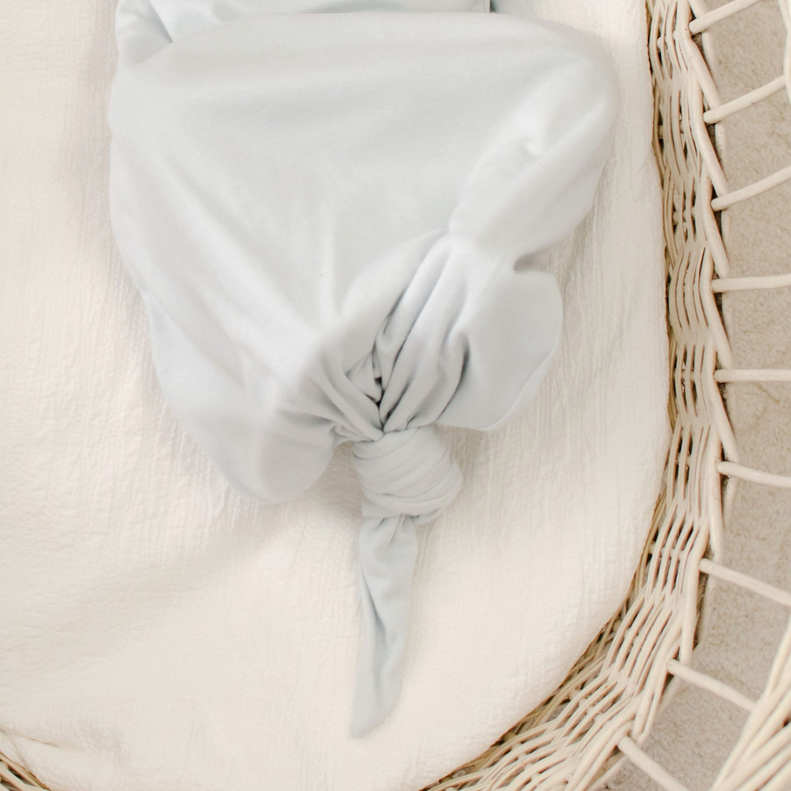 The Evan Knot Gown, made from 100% pima cotton in a light gray hue and handcrafted in the USA, is elegantly tied in a knot for a smooth, gathered look. Nestled inside a wicker basket on a textured white surface, this newborn boys' gown radiates softness and tranquility.