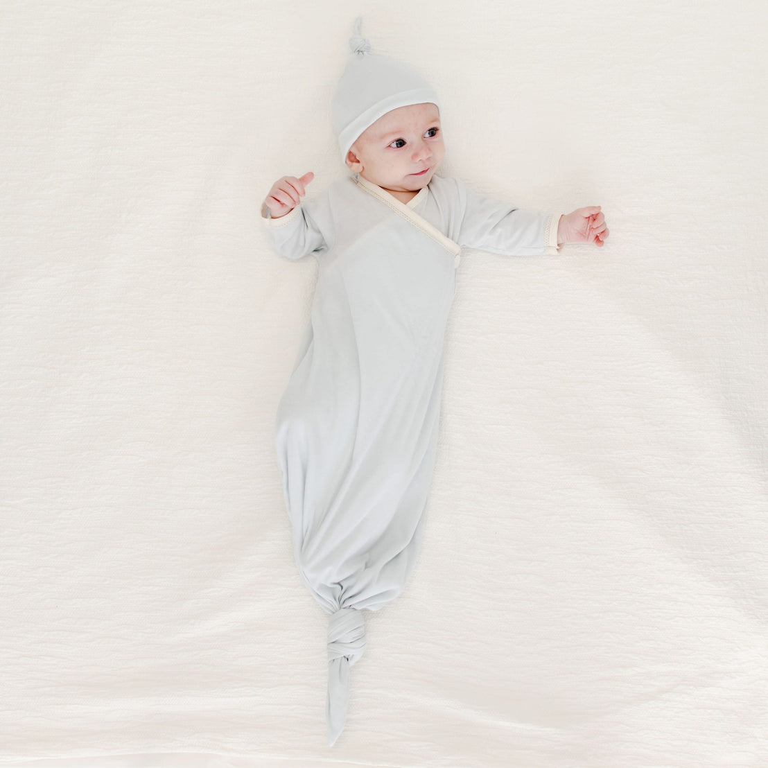 A baby in the Evan Knot Gown, artisanally crafted from 100% pima cotton, wears a coordinating hat while reclining on a cozy, cream-colored surface. The baby looks upward with arms gently outstretched. Skillfully made in the USA with love and care.