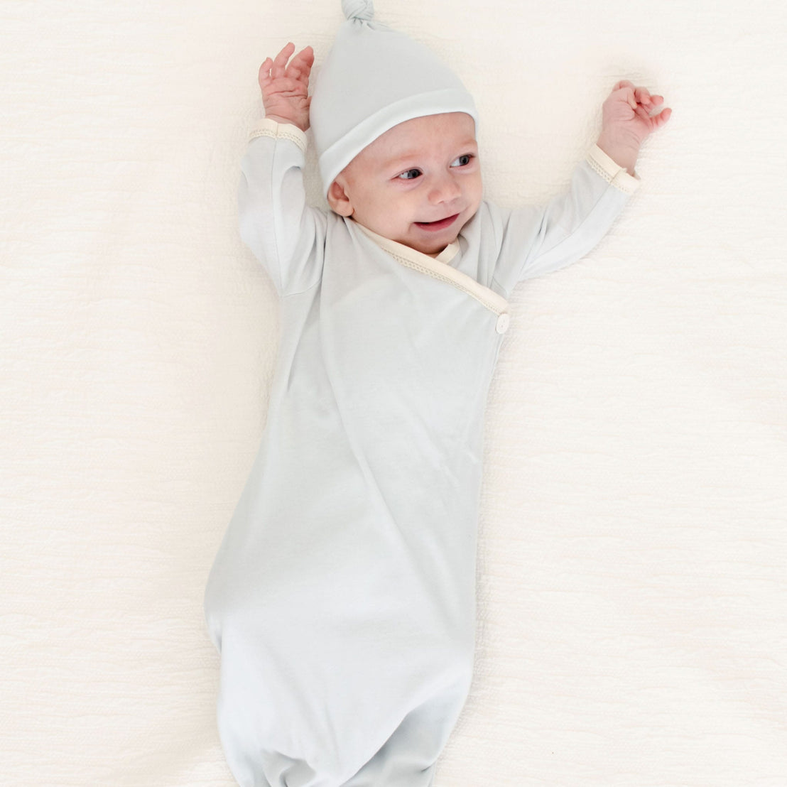 A newborn boy, dressed in the Evan Knot Gown with a matching hat, lies joyfully on a soft surface. This delightful outfit is expertly handmade in the USA from 100% pima cotton, ensuring both comfort and style as he stretches his arms above his head with a smile.