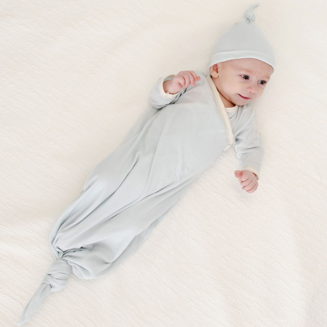 A baby lies on a soft white blanket, smiling and looking upward, dressed in the Evan Knot Gown—a light gray newborn boys outfit handmade in the USA from 100% pima cotton—with a matching hat.