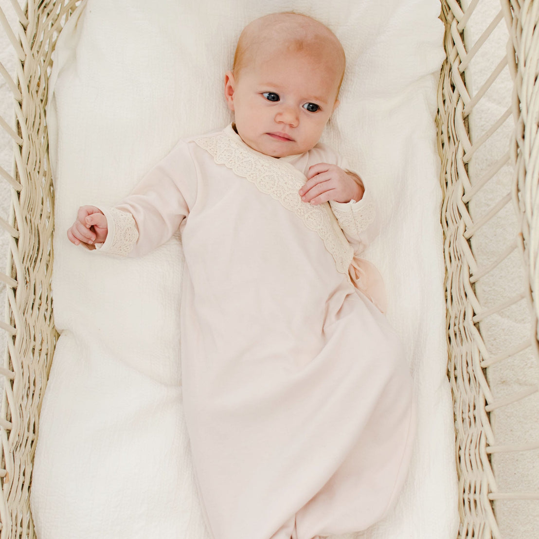 A newborn wearing the Evelyn Knot Gown with lace detailing lies in a white wicker basket, looking up with a calm expression.