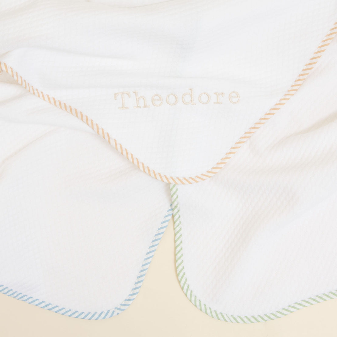 The First Easter Personalized Blanket is crafted from soft, white fabric and showcases custom embroidery of the name "Theodore" in the center. This charming gift features pastel-colored diagonal striped borders in blue, green, and orange hues, with a slightly crumpled texture adding to its appeal.