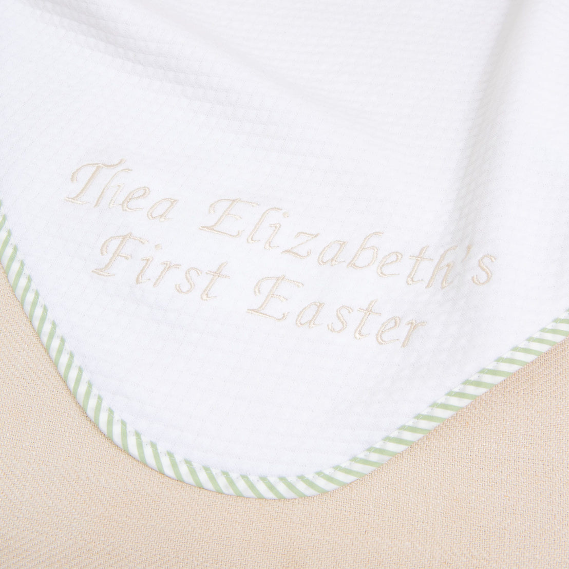 A white blanket featuring green striped edging and custom embroidery that reads "Thea Elizabeth's First Easter" in light brown thread makes it a perfect personalized gift. This is the First Easter Personalized Blanket.