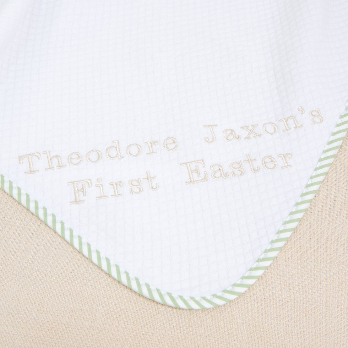 A close-up of the First Easter Personalized Blanket reveals a white fabric with green striped edging, customized with "Theodore Jaxon's First Easter" embroidered in light brown stitches. This personalized keepsake boasts a subtle grid pattern, ideal for celebrating a first Easter.