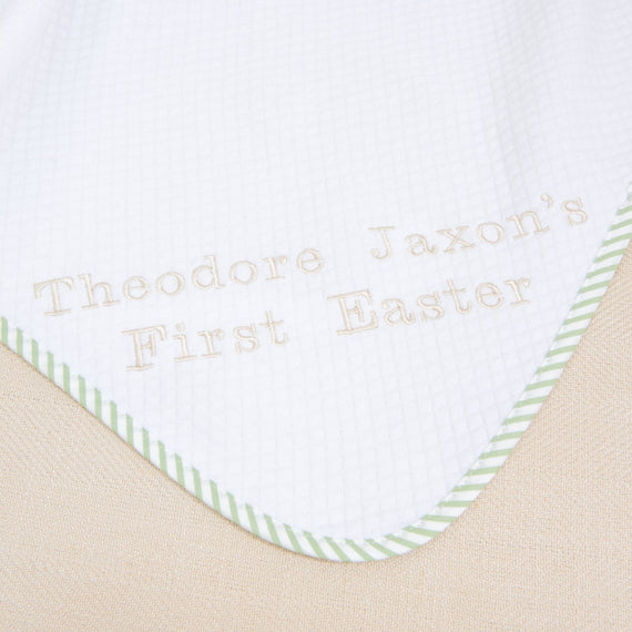 A close-up of the First Easter Personalized Blanket reveals a white fabric with green striped edging, customized with "Theodore Jaxon's First Easter" embroidered in light brown stitches. This personalized keepsake boasts a subtle grid pattern, ideal for celebrating a first Easter.