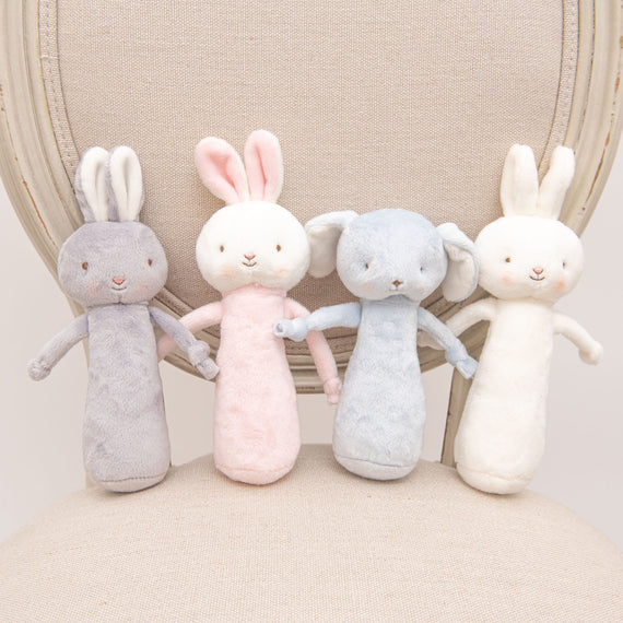 Four Friendly Chime Rattles in gray, pink, blue, and white are lined up on a beige chair with a round back. These baby-safe toys have elongated bodies and floppy ears made from soft, textured fabric featuring soft embroidered details for eyes and noses.