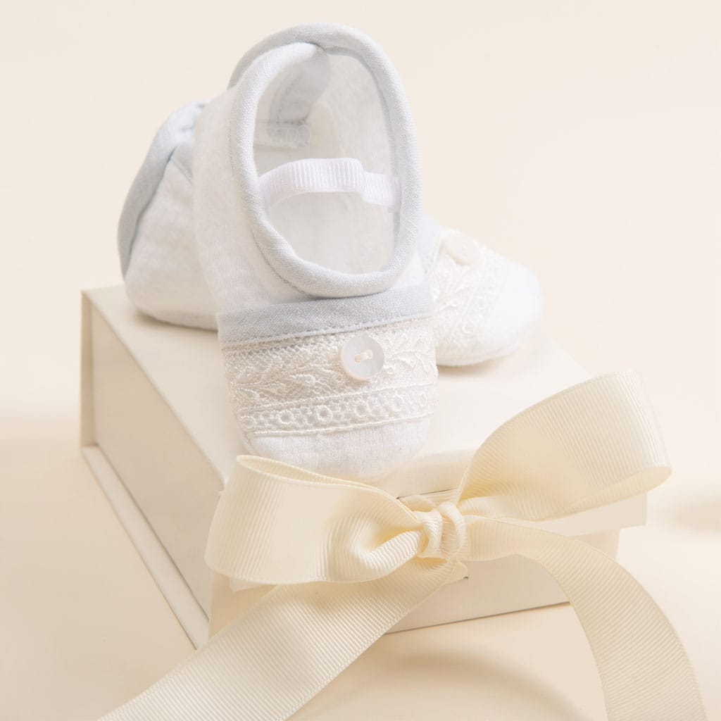 A pair of white baby shoes with lace and button details sits on the gift wrap titled "Mini Gift Box with Bow" from baby beau & belle, featuring an ivory tissue and cream-colored ribbon bow. The background is a soft, neutral color.