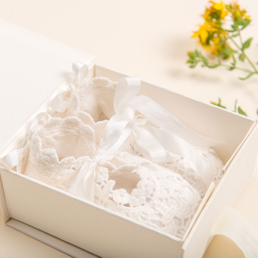 A pair of delicate white baby booties featuring lace and ribbon embellishments is elegantly presented in an open cream-colored mini gift box, adorned with a soft ivory tissue lining. A small yellow flower in the background adds a touch of color, enhancing this charming scene by Baby Beau & Belle.