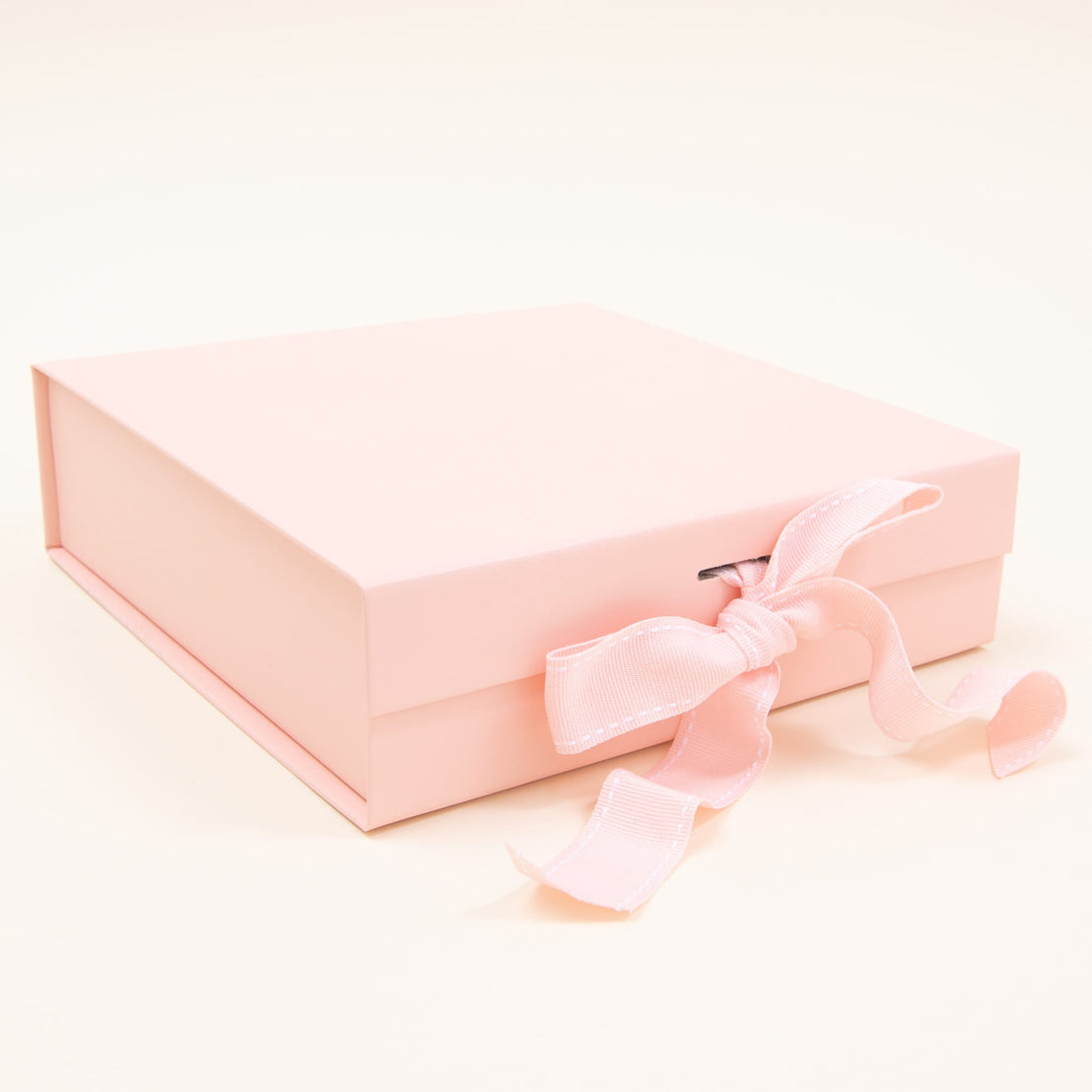 A closed rectangular gift box by Baby Beau & Belle, finished in a pale pink hue with a matching satin ribbon tied into a bow on the side, is beautifully presented against a simple light background.