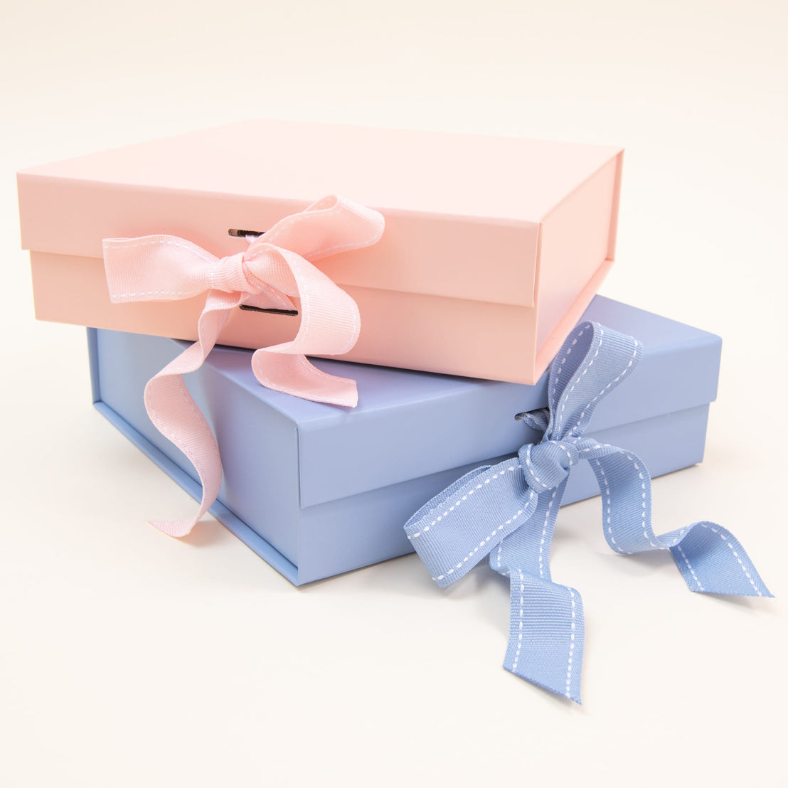 Two sophisticated "Gift Wrap | Small Gift Box with Bow" packages are artistically arranged against a soft backdrop. A pink box adorned with a delicate pale pink ribbon pairs beautifully with a blue box, which is accented by a blue ribbon featuring white stitching. Ideal for any occasion, these charming boxes embody the elegance of Baby Beau & Belle.