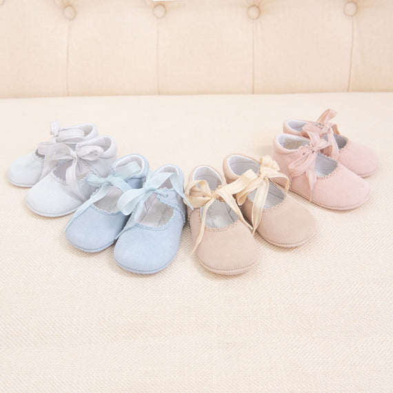 Three pairs of Girls Suede Tie Mary Janes in blue, beige, and pink, each tied with a ribbon, arranged in a row on a cream background with a tufted cushion behind.