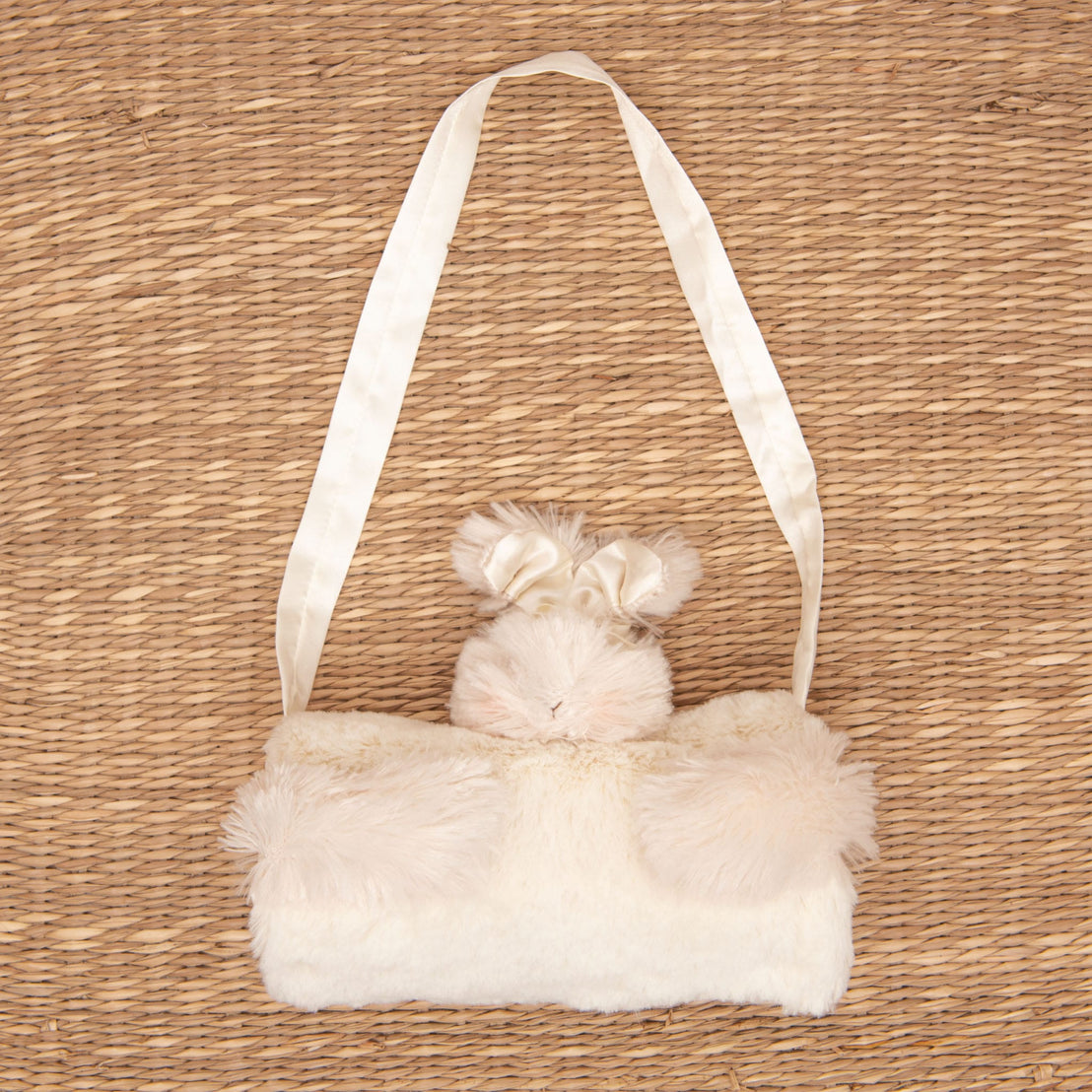 The Glad Dreams Coat Set, featuring a plush and furry cream-colored handbag adorned with a bunny design complete with ears, rests on a woven straw surface. Resembling a faux fur baby coat, it includes a satin strap and soft fur accents, offering a cute and cozy style. This set is perfect for those seeking a unique baby gift.